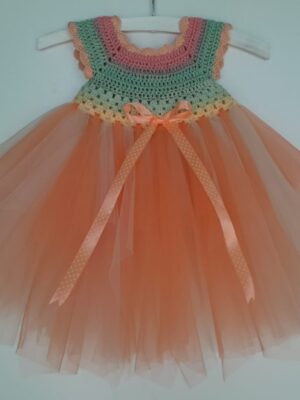 A baby wearing a rainbow crocheted bodice with a peach tulle skirt. Their hands trace the satin ribbon under the bodice.
